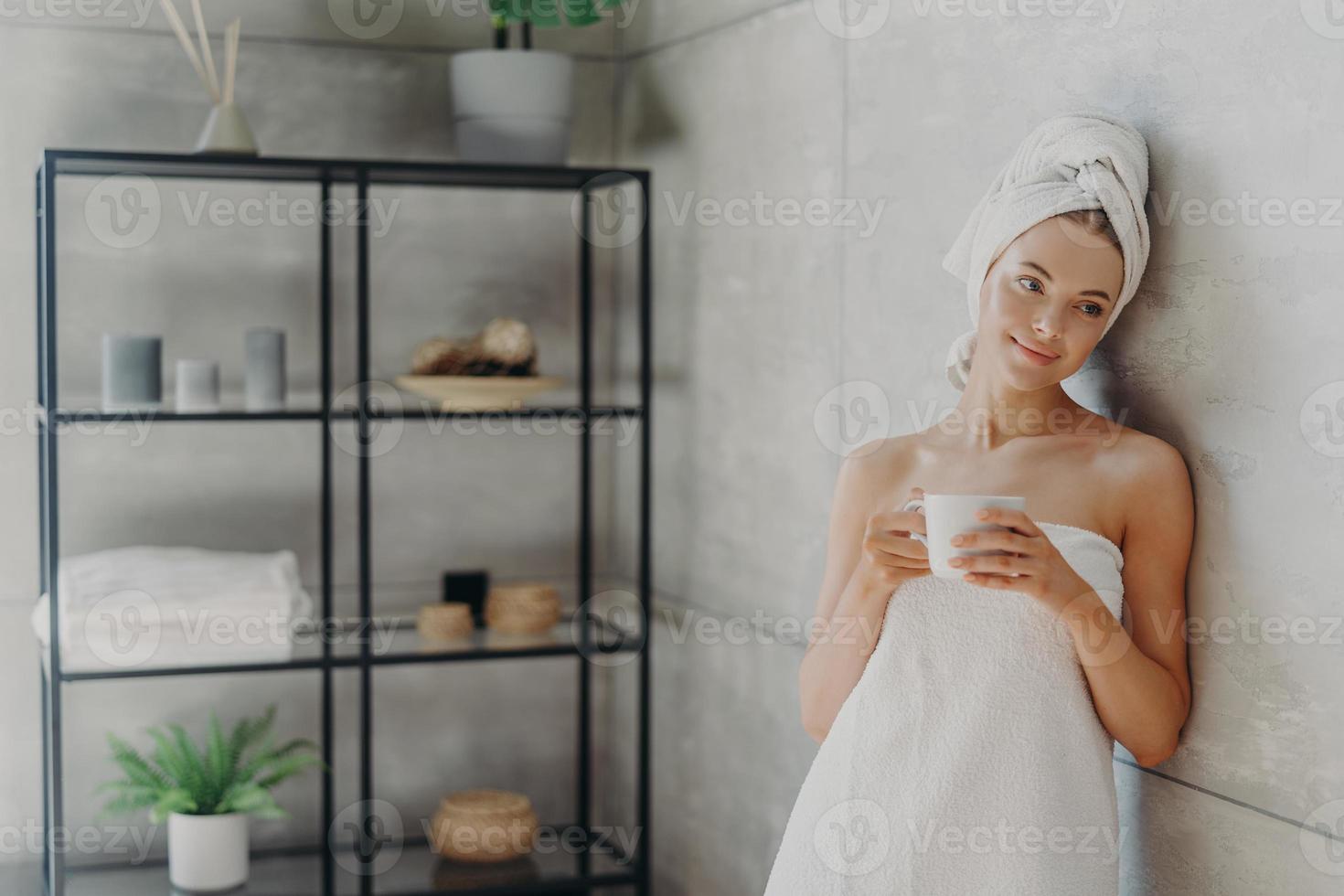 foto van rustige ontspannen vrouw poseert in handdoek tegen badkamer interieur, gewikkeld in badhanddoek, drinkt thee of koffie, heeft een schone perfecte huid, leunt tegen grijze muur. schoonheidsdag, spa en huidverzorging