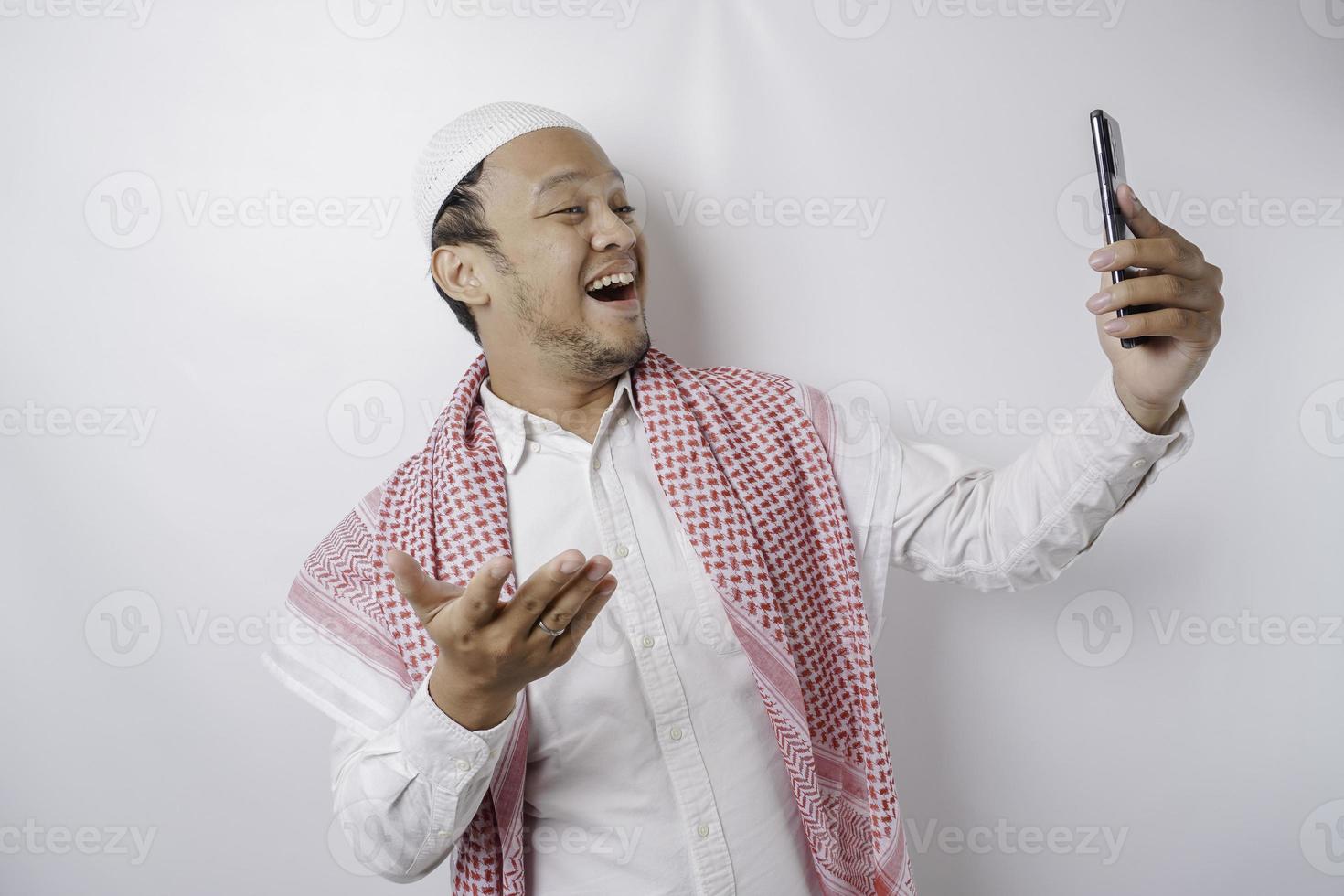 een portret van een gelukkig Aziatisch moslim Mens glimlachen terwijl Holding zijn telefoon, geïsoleerd door wit achtergrond foto