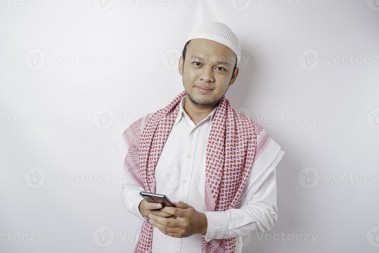 een portret van een gelukkig Aziatisch moslim Mens glimlachen terwijl Holding zijn telefoon, geïsoleerd door wit achtergrond foto