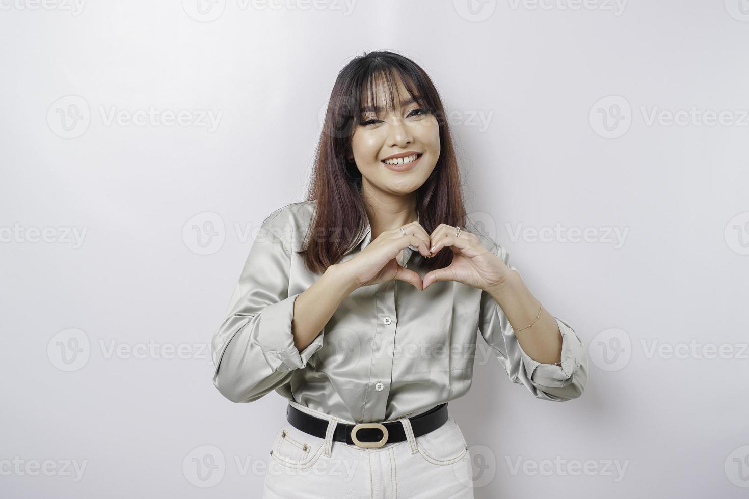 een aantrekkelijk jong Aziatisch vrouw vervelend een salie groen overhemd voelt gelukkig en een romantisch vormen hart gebaar drukt uit inschrijving gevoelens foto
