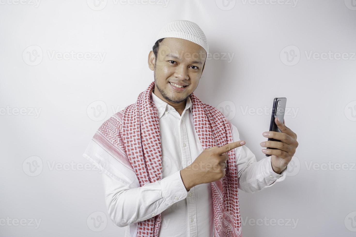 een portret van een gelukkig Aziatisch moslim Mens glimlachen terwijl Holding zijn telefoon, geïsoleerd door wit achtergrond foto