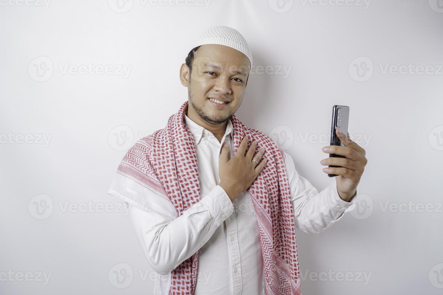 een portret van een gelukkig Aziatisch moslim Mens glimlachen terwijl Holding zijn telefoon, geïsoleerd door wit achtergrond foto