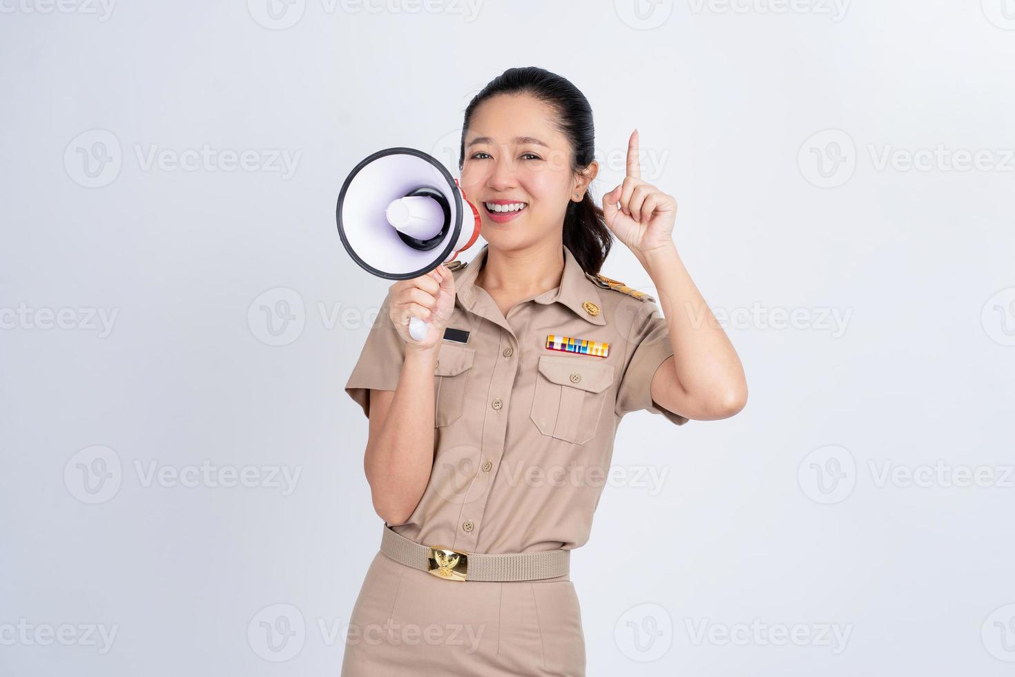 gelukkig jong Aziatisch vrouw in bruin uniform werk kleren Holding megafoon geïsoleerd Aan wit achtergrond, ze was richten omhoog met haar hand. foto