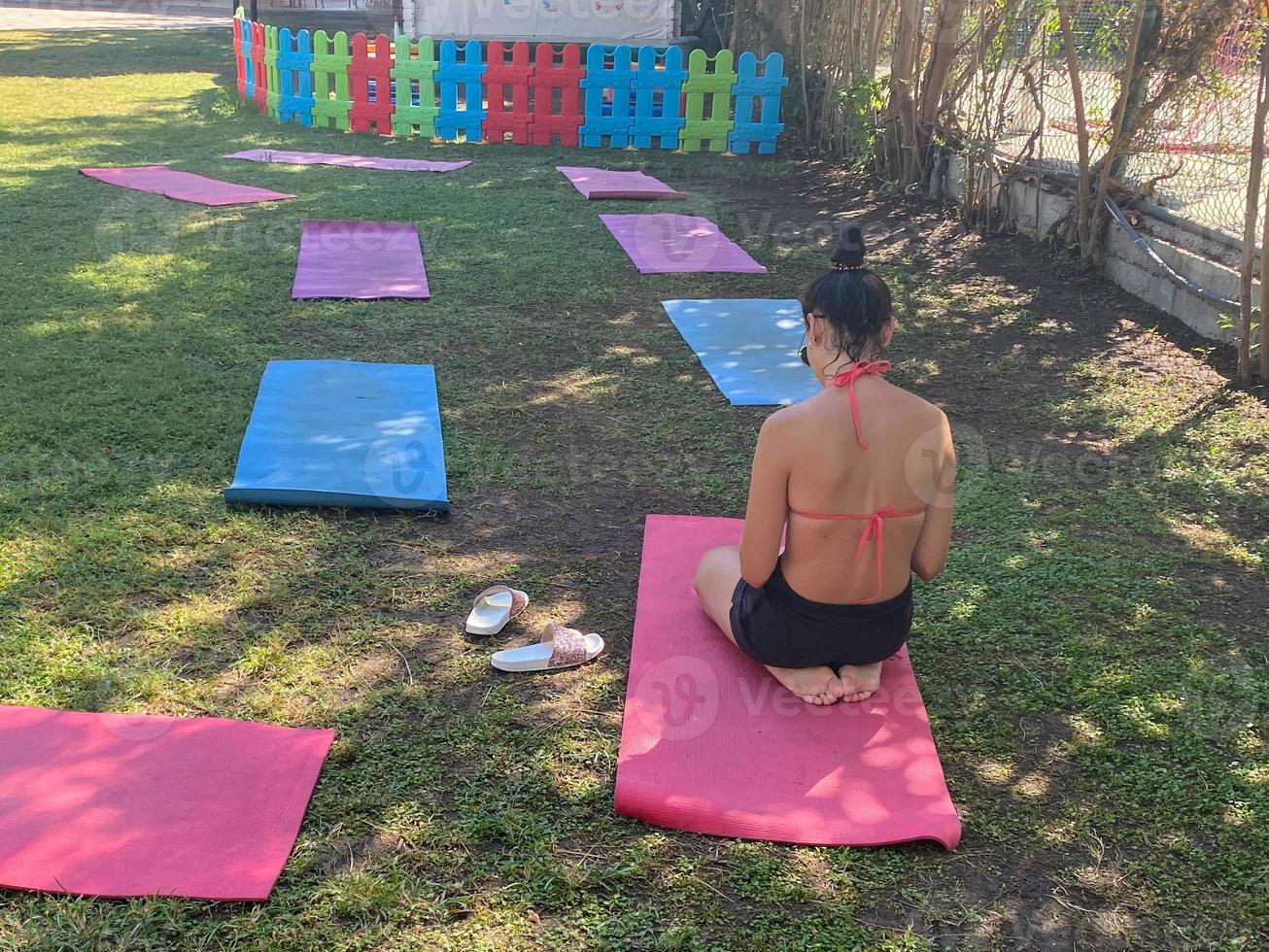 hardwerkend, zoet, peuter- meisje, kind atleet zit Aan een groen tapijt in een lotus positie, poten gekruist, het uitvoeren van sport- opdrachten, yoga en gymnastiek, uitrekken haar armen foto