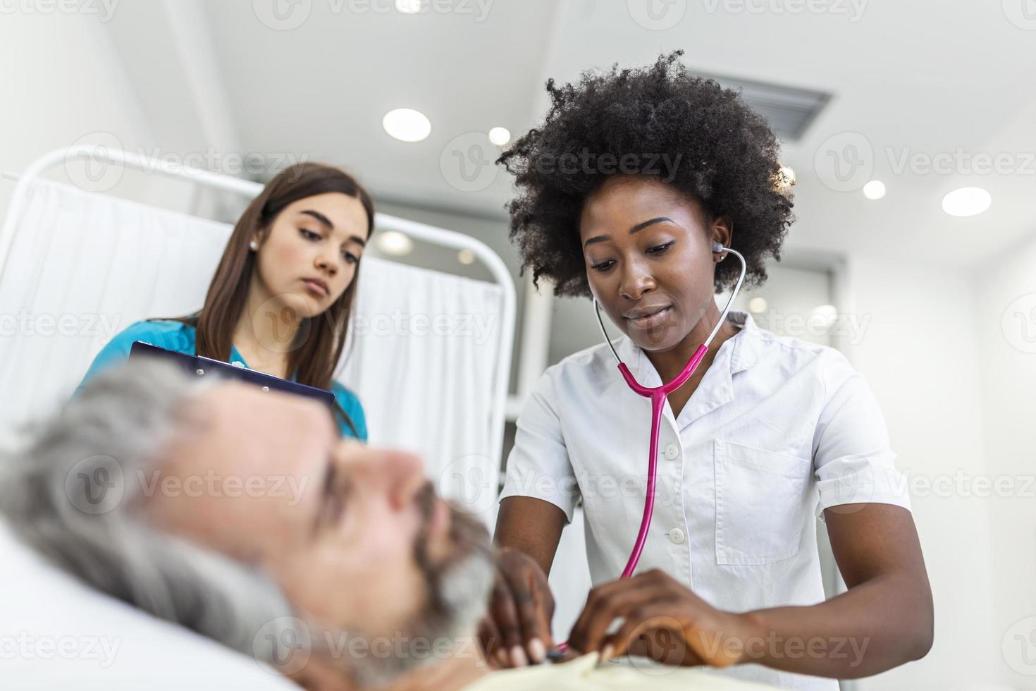 ziek Mens geduldig met Afrikaanse Amerikaans vrouw dokter luistert zijn borst met stethoscoop in ziekenhuis noodgeval kamer. dokter en verpleegster komen naar bezoek ouderling geduldig Bij ziekenhuis kamer. foto