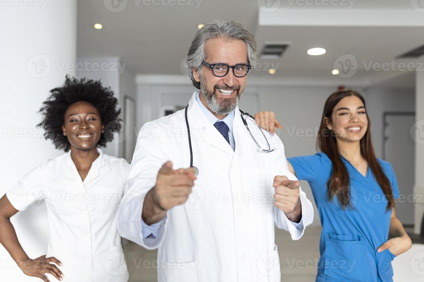 een multi-etnisch groep van drie artsen en verpleegsters staand in een ziekenhuis hal, vervelend scrubs en jassen. de team van gezondheidszorg arbeiders zijn staren Bij de camera en glimlachen foto