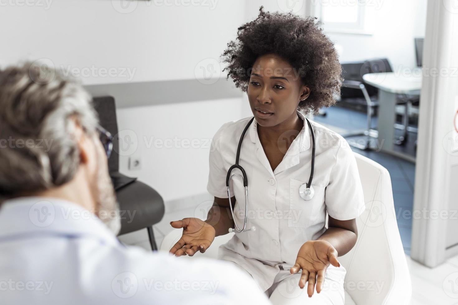 zwart vrouw dokter en senior Mens communiceren in een aan het wachten kamer Bij ziekenhuis. geneesmiddel, gezondheidszorg en mensen concept - dokter en geduldig vergadering Bij ziekenhuis foto