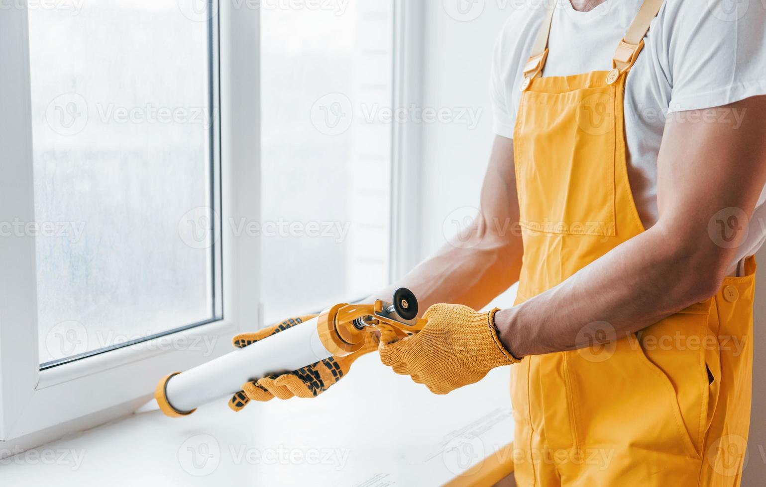 klusjesman in geel uniform werken met lijm voor venster binnenshuis. huis vernieuwing opvatting foto
