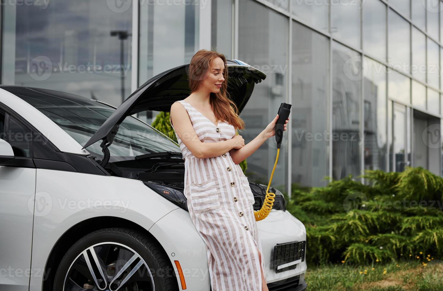 looks Bij apparaat. vrouw Aan de elektrisch auto's in rekening brengen station Bij dag. merk nieuw voertuig foto