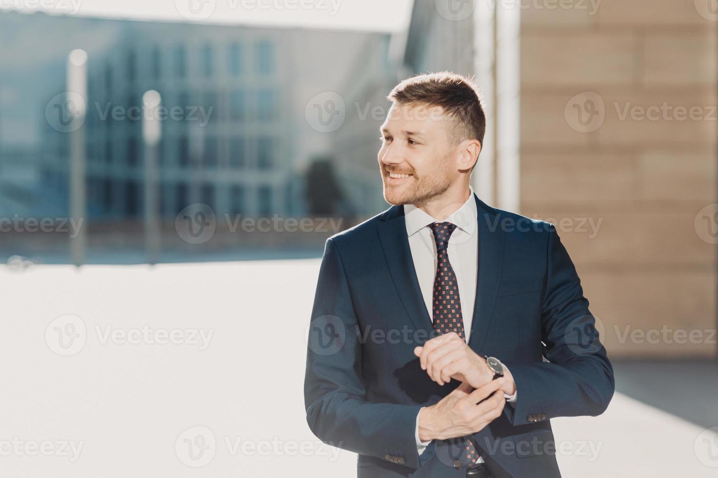 mensen en bedrijfsconcept. blije elegante jonge zakenman met stoppels, draagt formele kleding en horloge, wacht op iemand, poseert buiten in de buurt van kantoor. mensen, carrière en leiderschap concept. foto