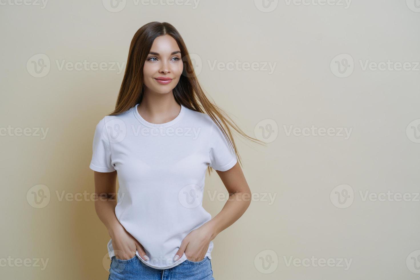 nadenkende brunette vrouw kijkt opzij, draagt een casual wit t-shirt, houdt de handen in de zakken, kijkt opzij, geïsoleerd over beige achtergrond, heeft een ontspannen gezichtsuitdrukking, lege ruimte voor uw promotie foto