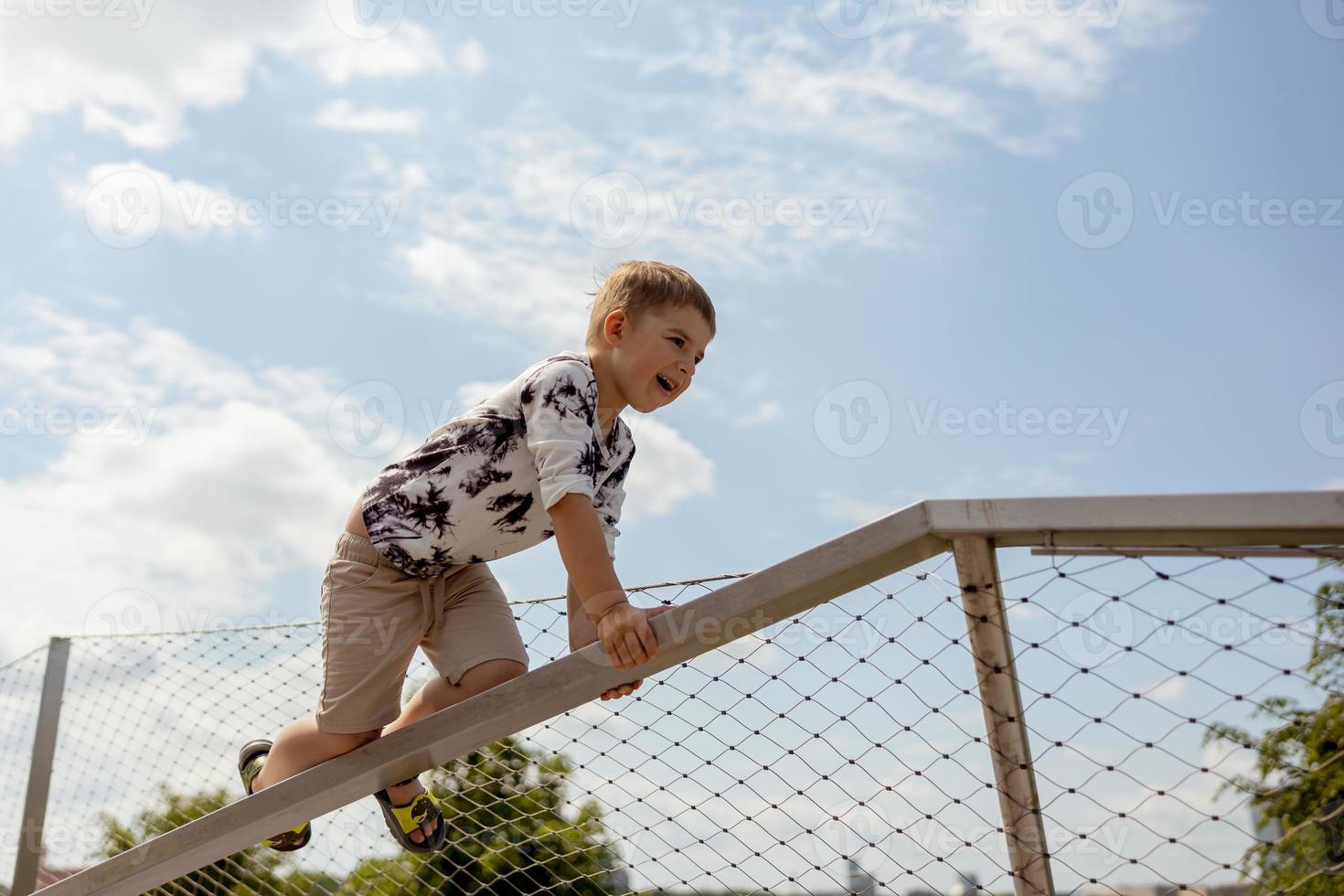 aanbiddelijk Kaukasisch jongen beklimming Aan metaal leuning buitenshuis. schattig kind hebben pret in de stad. actief vrije tijd. mooi visie, blauw lucht, verbazingwekkend zonnig het weer. Gaan naar voren, beklimmen naar de top. foto