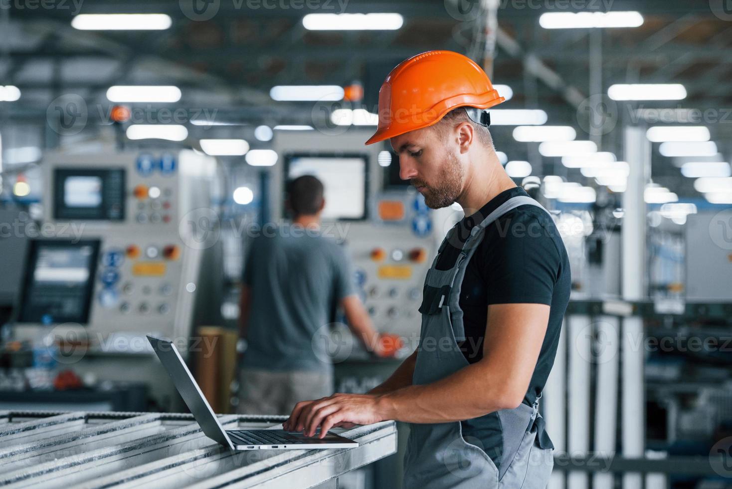 modern laptop. typen Aan toetsenbord. industrieel arbeider binnenshuis in fabriek. jong technicus met oranje moeilijk hoed foto