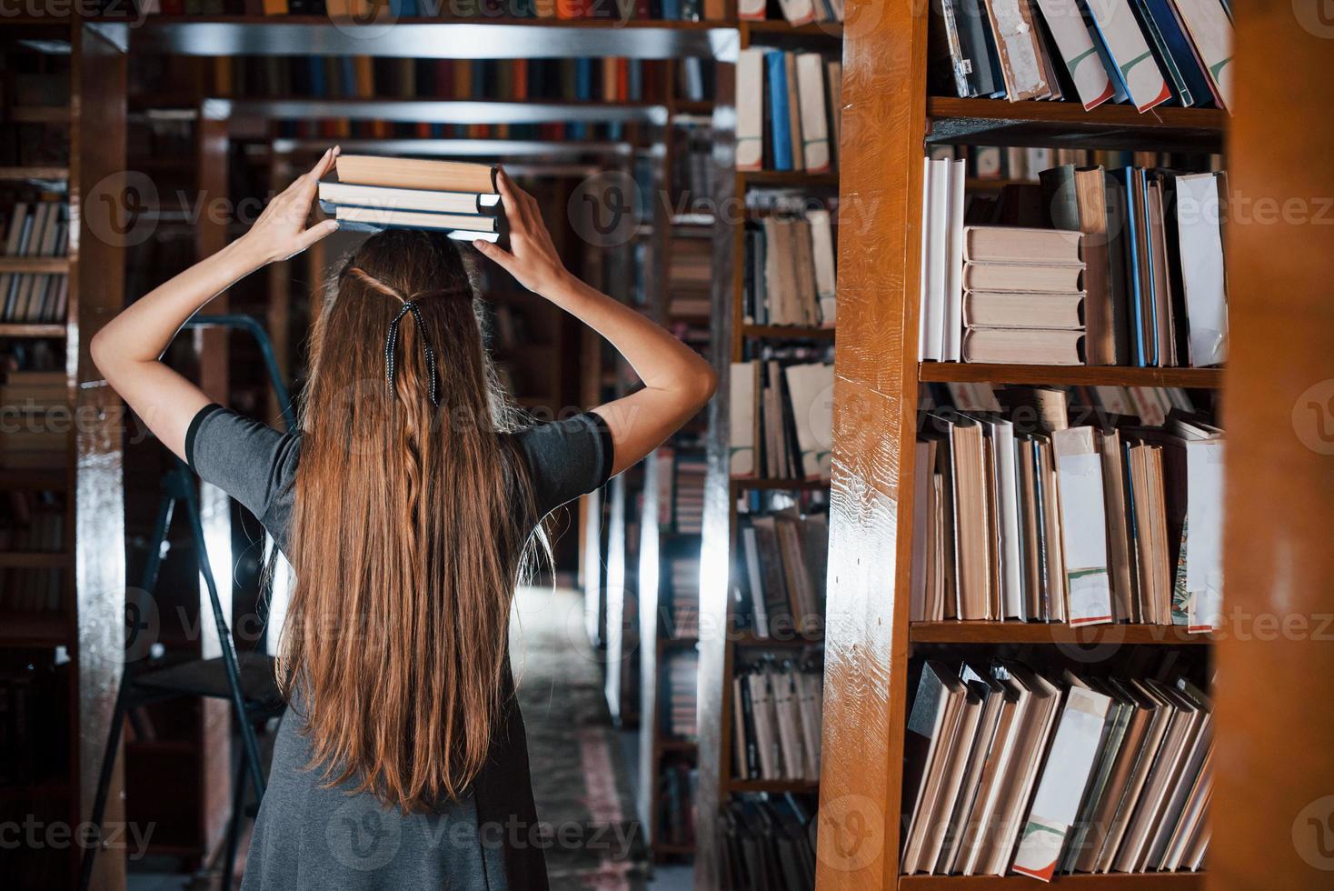 vrolijk meisje hebben pret terwijl zet boeken Aan haar hoofd. vrouw leerling is in bibliotheek. opvatting van onderwijs foto