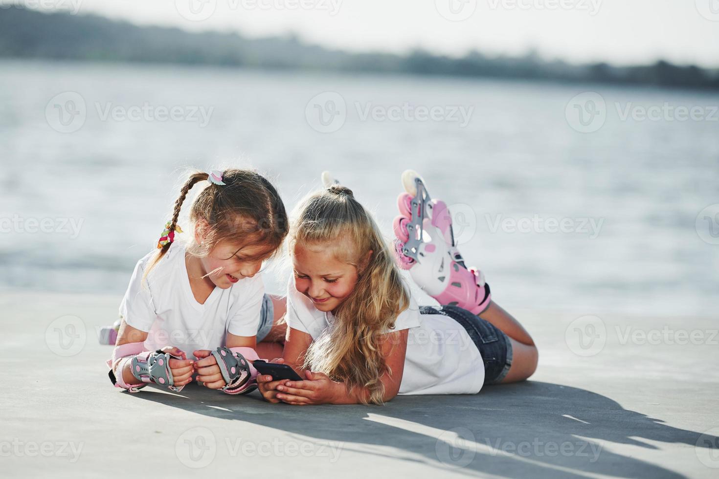 aan het liegen omlaag. twee weinig meisjes met rol skates buitenshuis in de buurt de meer Bij achtergrond foto