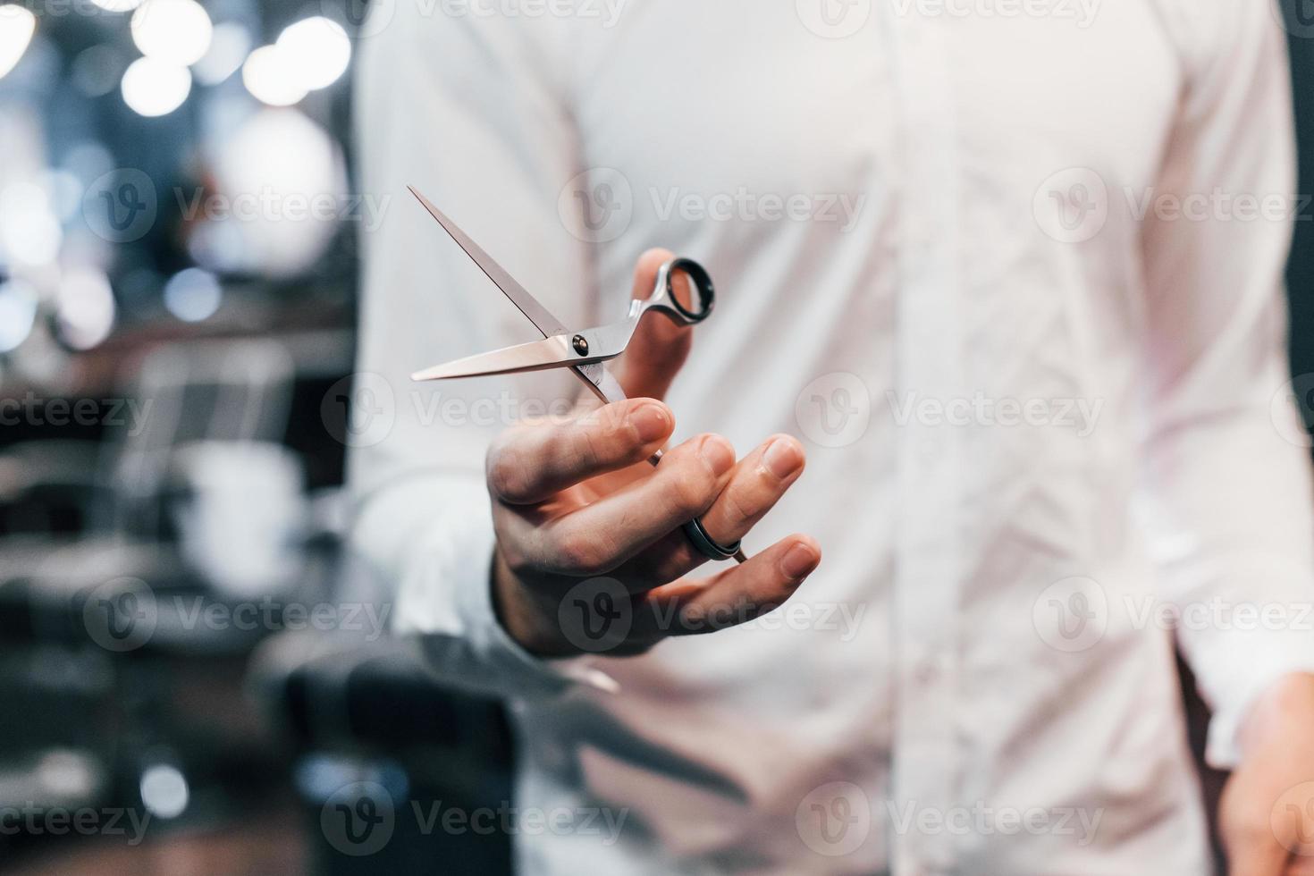 dichtbij omhoog visie van vent in wit kleren dat staand binnenshuis in kapper winkel en Holding schaar foto