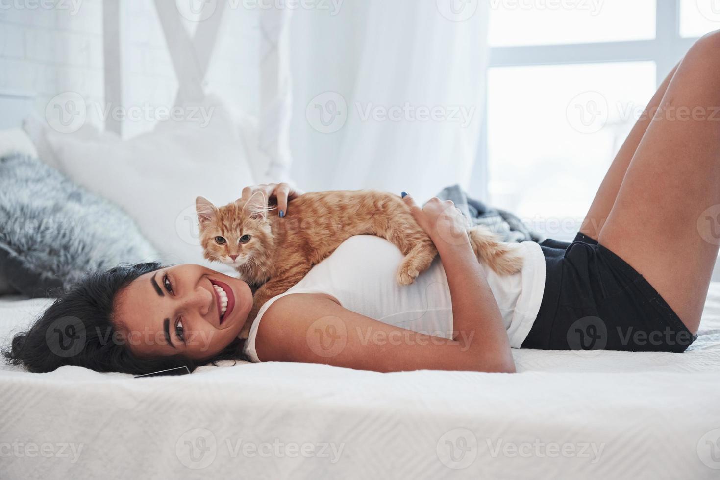 kat aan het liegen Aan de meisje. aantrekkelijk blond resting Aan de wit bed met haar schattig katje foto