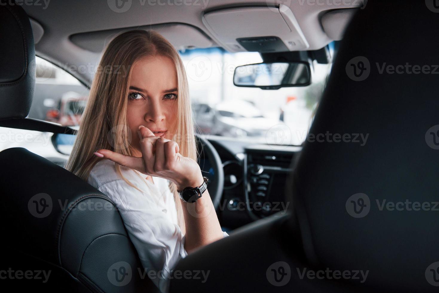 draaide zich om terug naar de cameramannen Bij achterbank. mooi blond meisje zittend in de nieuw auto met modern zwart interieur foto