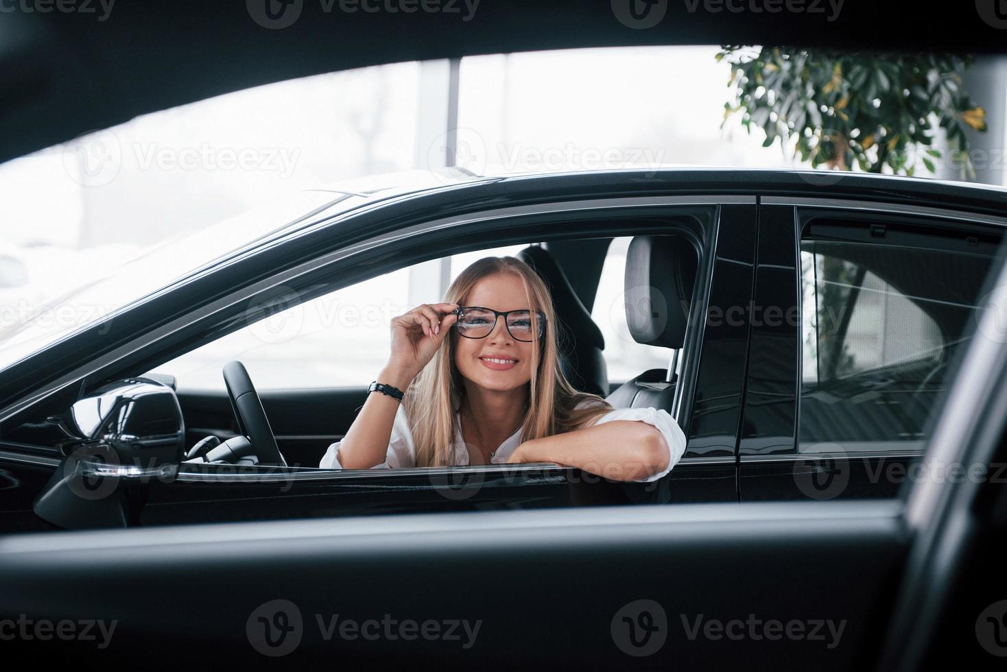 foto door de geopend ramen. mooi blond meisje zittend in de nieuw auto met modern zwart interieur