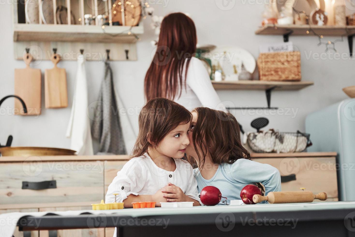 kinderen zijn pratend heimelijk naar elk andere in de keuken terwijl mam is voorbereidingen treffen avondeten foto