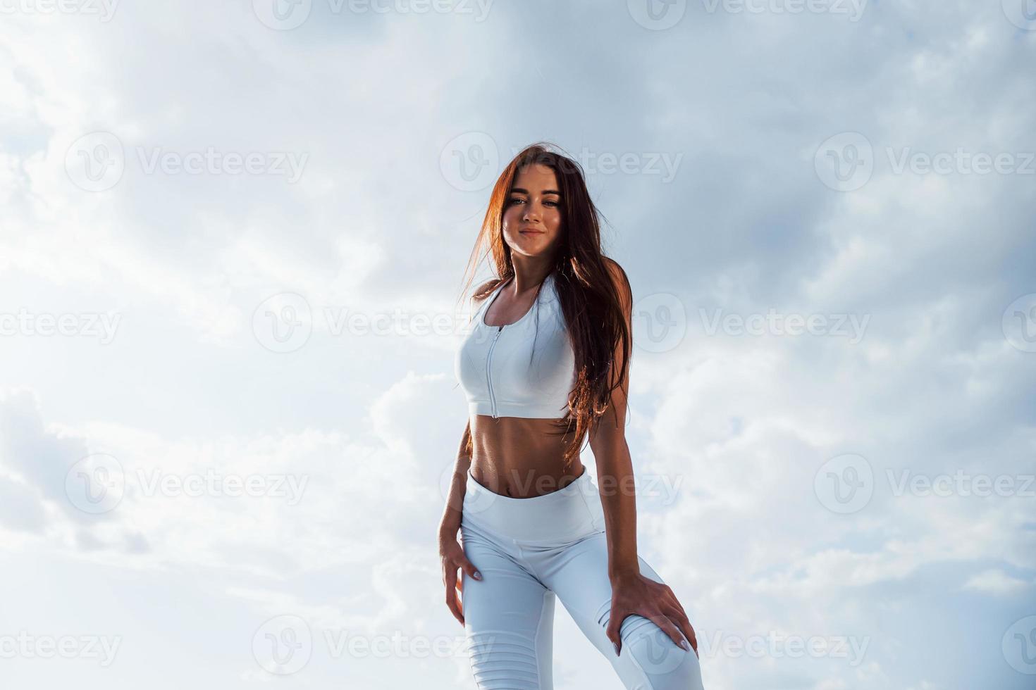 brunette geschiktheid vrouw staat buitenshuis in wit sportief kleren tegen bewolkt lucht Bij dag foto