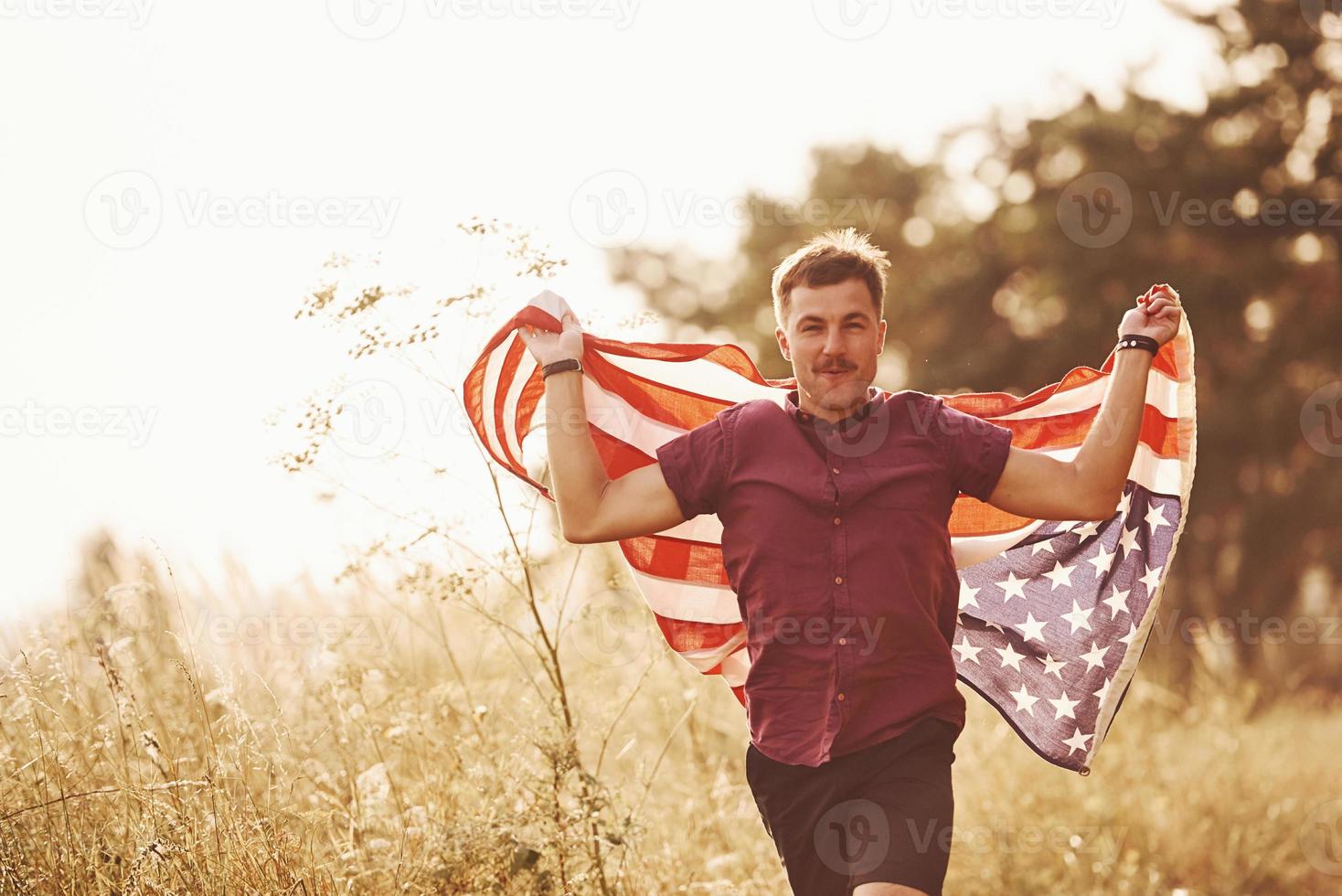 volwassen Mens loopt met Amerikaans vlag in handen buitenshuis in de veld. voelt vrijheid Bij zonnig dag foto