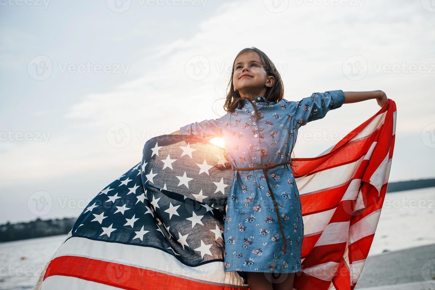 patriottisch vrouw kind met Amerikaans vlag in handen. tegen bewolkt lucht foto