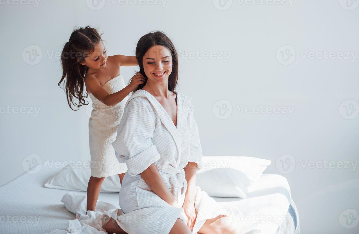 helpen naar gemaakt kapsel. jong moeder met haar dochter hebben schoonheid dag binnenshuis in wit kamer foto