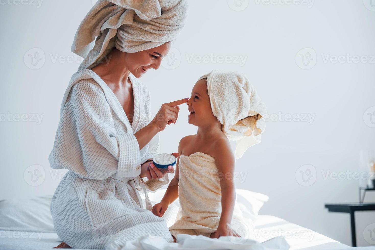 gebruik makend van room naar Doorzichtig huid. jong moeder met haar dochter hebben schoonheid dag binnenshuis in wit kamer foto