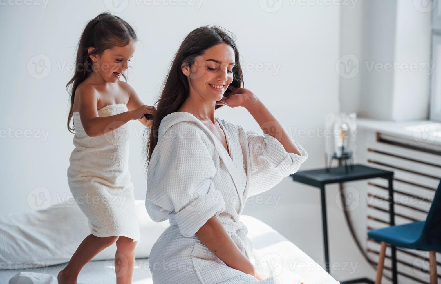 helpen naar gemaakt kapsel. jong moeder met haar dochter hebben schoonheid dag binnenshuis in wit kamer foto