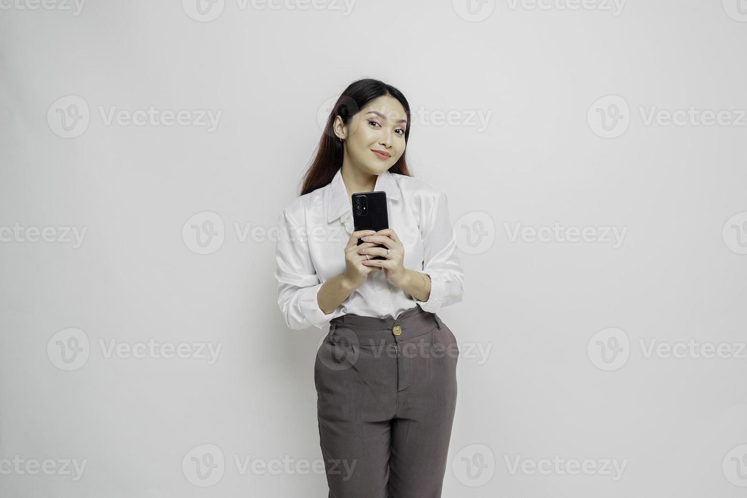 een portret van een gelukkig Aziatisch vrouw vervelend een wit overhemd en Holding haar telefoon, geïsoleerd door wit achtergrond foto
