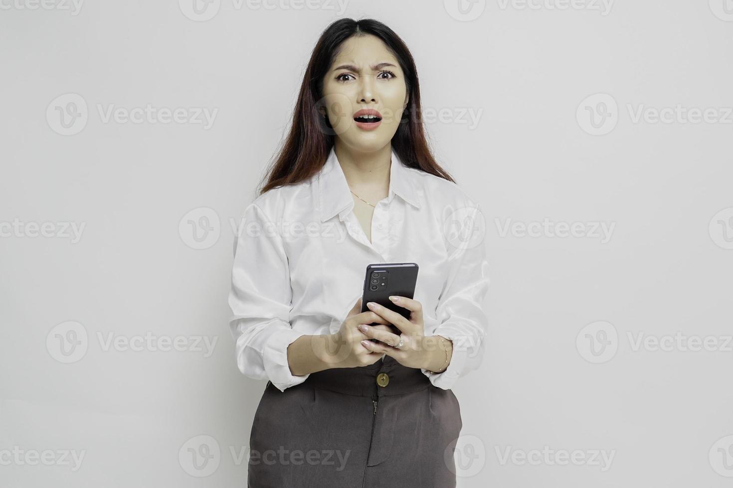 een ontevreden jong Aziatisch vrouw looks ontevreden vervelend wit overhemd geërgerd gezicht uitdrukkingen Holding haar telefoon foto