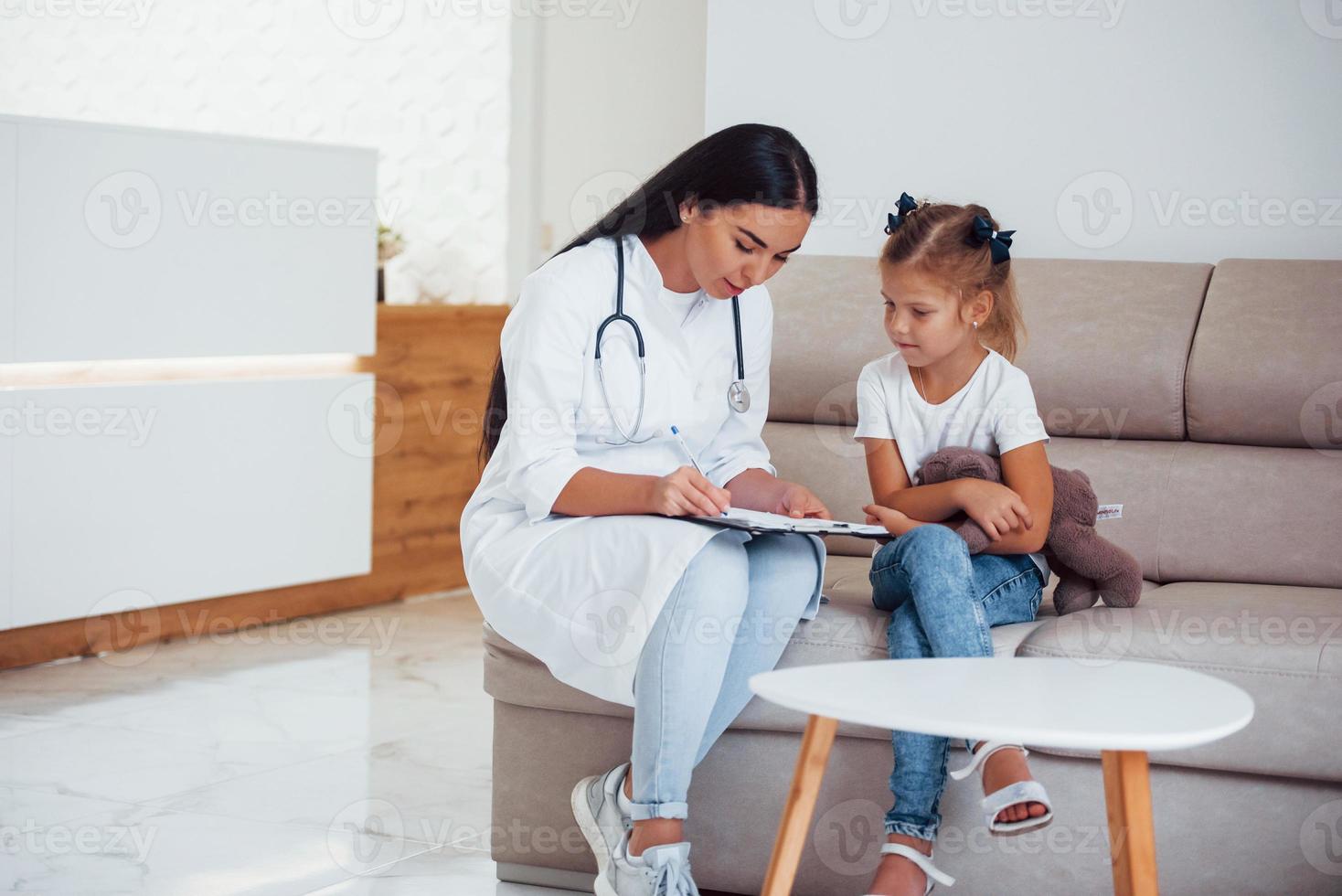 vrouw dokter met kladblok zit in de kliniek met weinig meisje foto