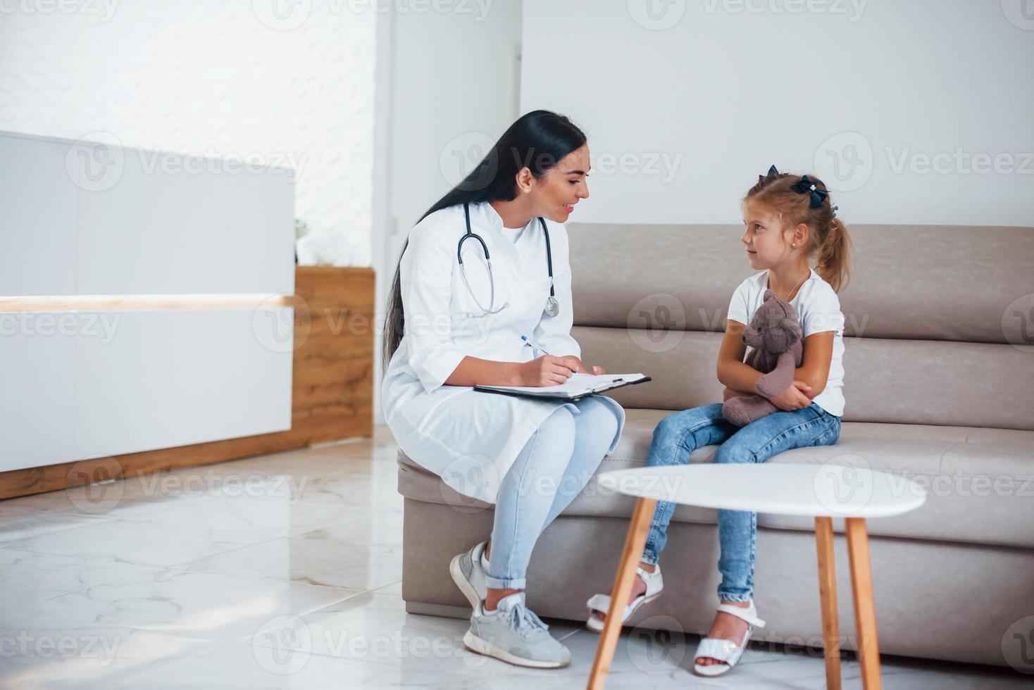 vrouw dokter met kladblok zit in de kliniek met weinig meisje foto