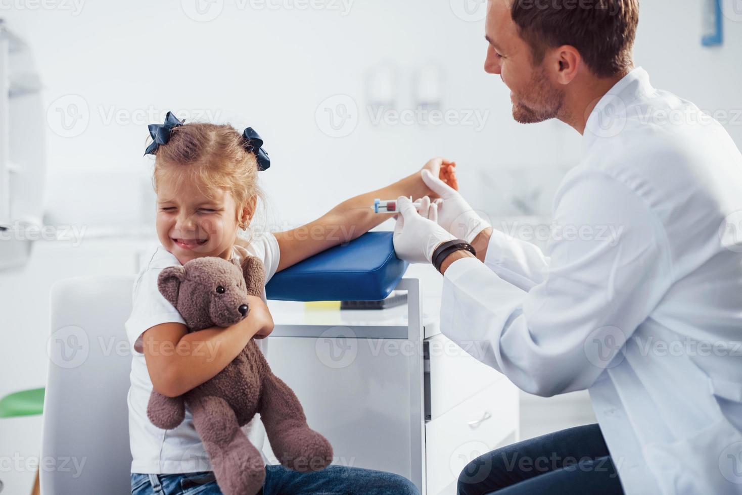 bloed bemonstering. weinig meisje met haar teddy beer is in de kliniek met dokter foto