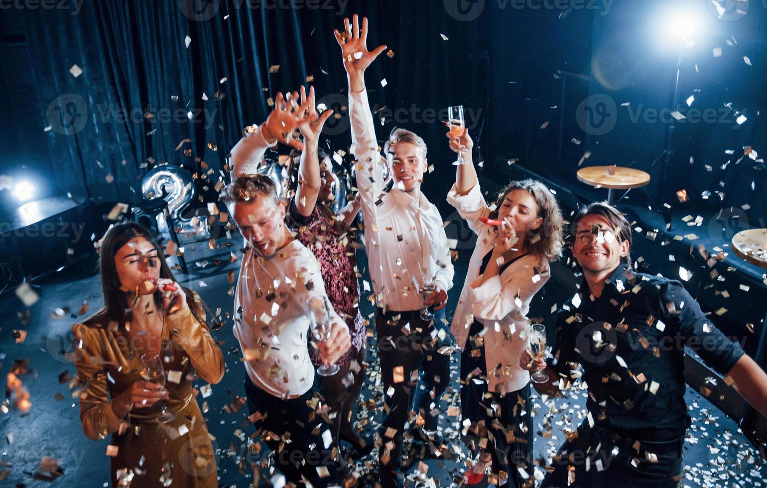 confetti is in de lucht. groep van vrolijk vrienden vieren nieuw jaar binnenshuis met drankjes in handen foto