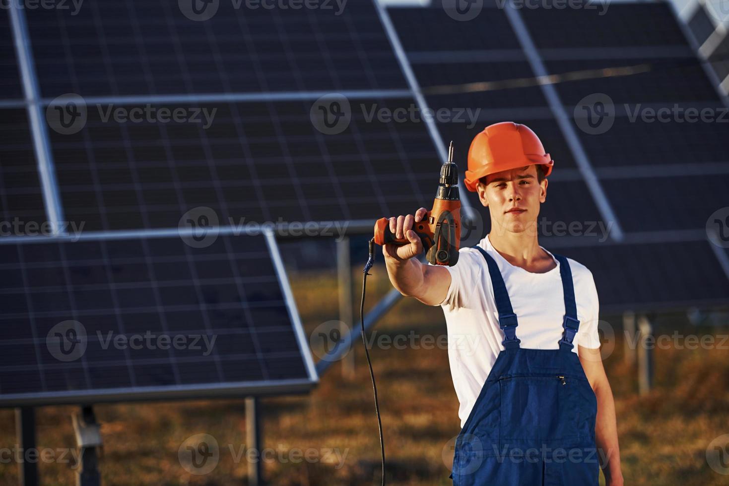 draadloos schroevedraaier in hand. mannetje arbeider in blauw uniform buitenshuis met zonne- batterijen Bij zonnig dag foto