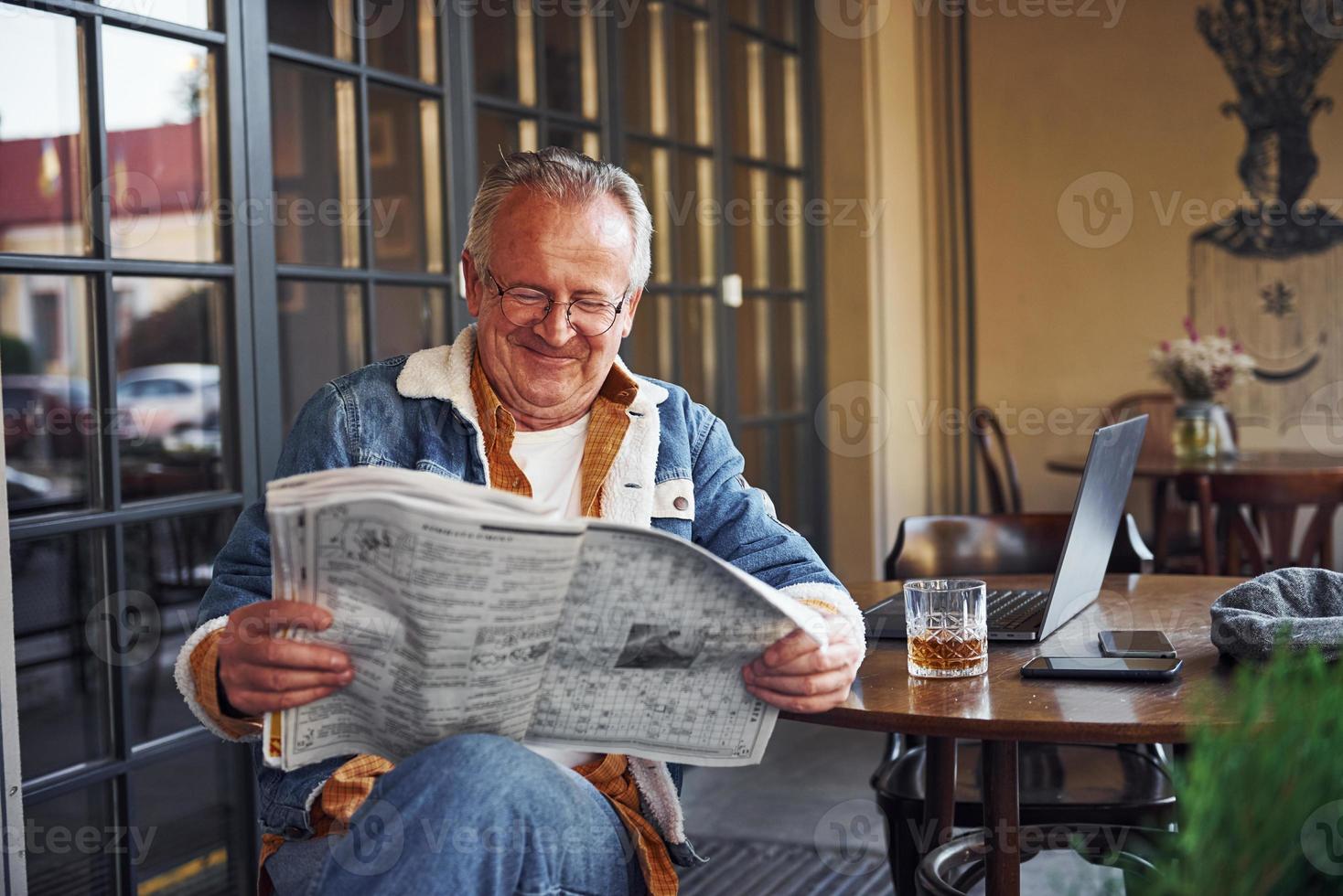 elegant senior in modieus kleren en in bril zit in de cafe en leest krant- foto