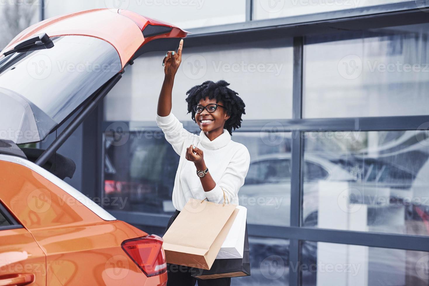 jong Afrikaanse Amerikaans vrouw in bril staat buitenshuis in de buurt modern auto na boodschappen doen met pakketjes foto