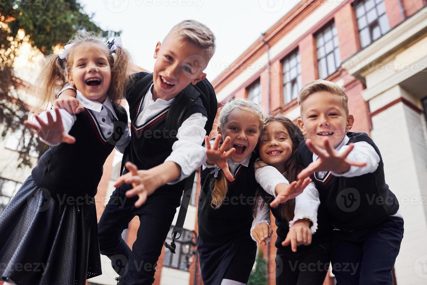 hebben pret en omarmen elk ander. groep van kinderen in school- uniform dat is buitenshuis samen in de buurt onderwijs gebouw foto