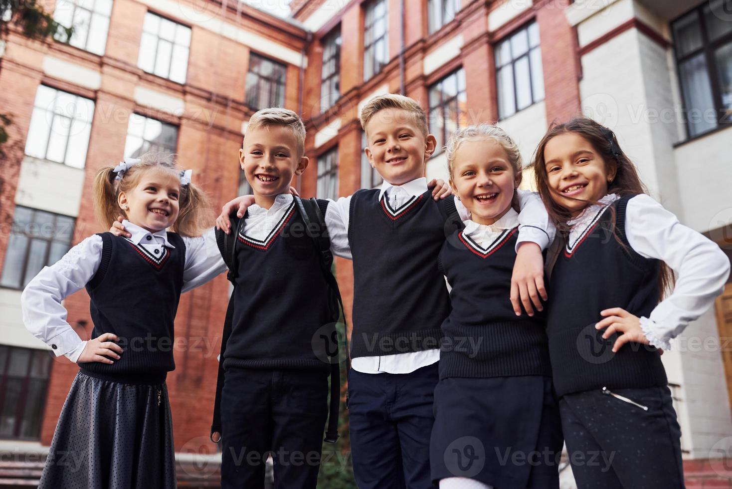 hebben pret en omarmen elk ander. groep van kinderen in school- uniform dat is buitenshuis samen in de buurt onderwijs gebouw foto