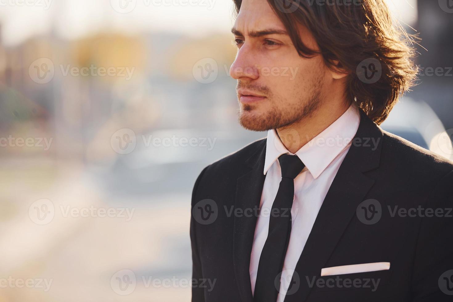 portret van knap jong zakenman in zwart pak en stropdas staat in de stad Bij zonnig dag foto