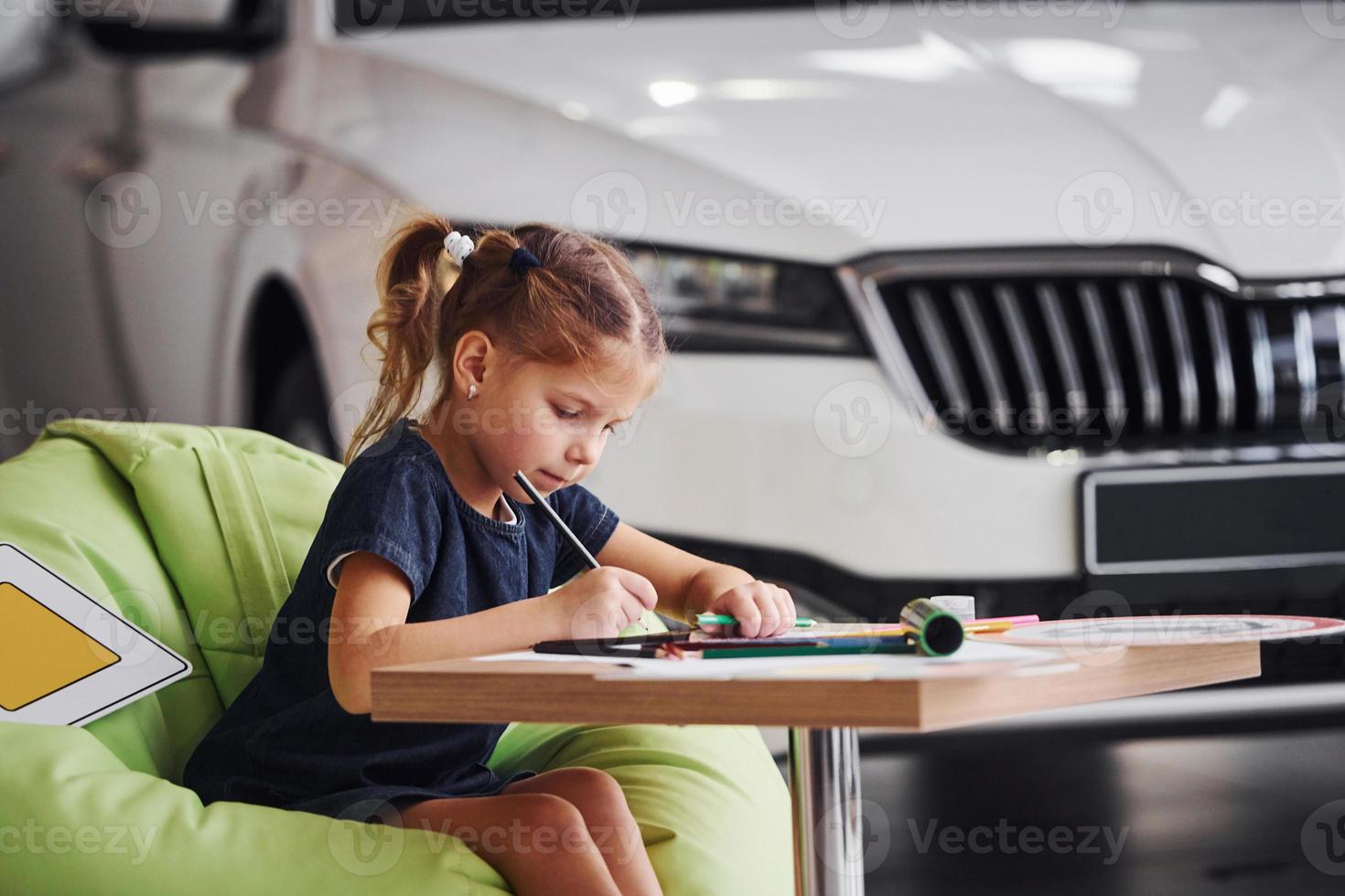 schattig weinig meisje zit Aan de zacht groen stoel door de tafel met potlood en papier bladen. in de buurt modern auto- foto