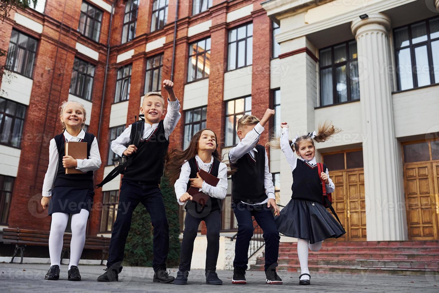 groep van kinderen in school- uniform jumping en hebben pret buitenshuis samen in de buurt onderwijs gebouw foto