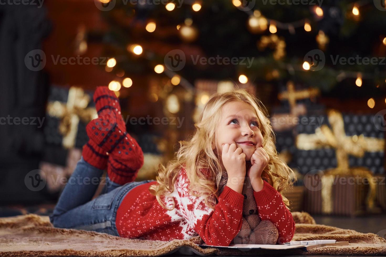 schattig weinig meisje in rood feestelijk trui aan het liegen naar beneden met teddy beer binnenshuis vieren nieuw jaar foto