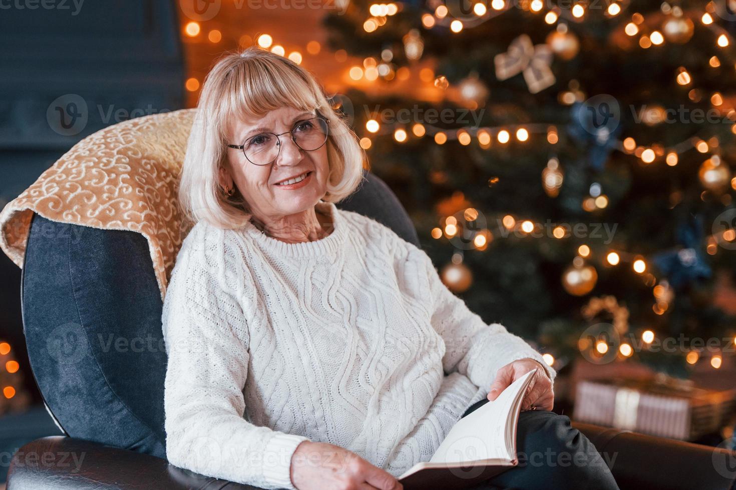 grootmoeder zittend Aan de stoel met boek in feestelijk nieuw jaar kamer met Kerstmis decoraties foto