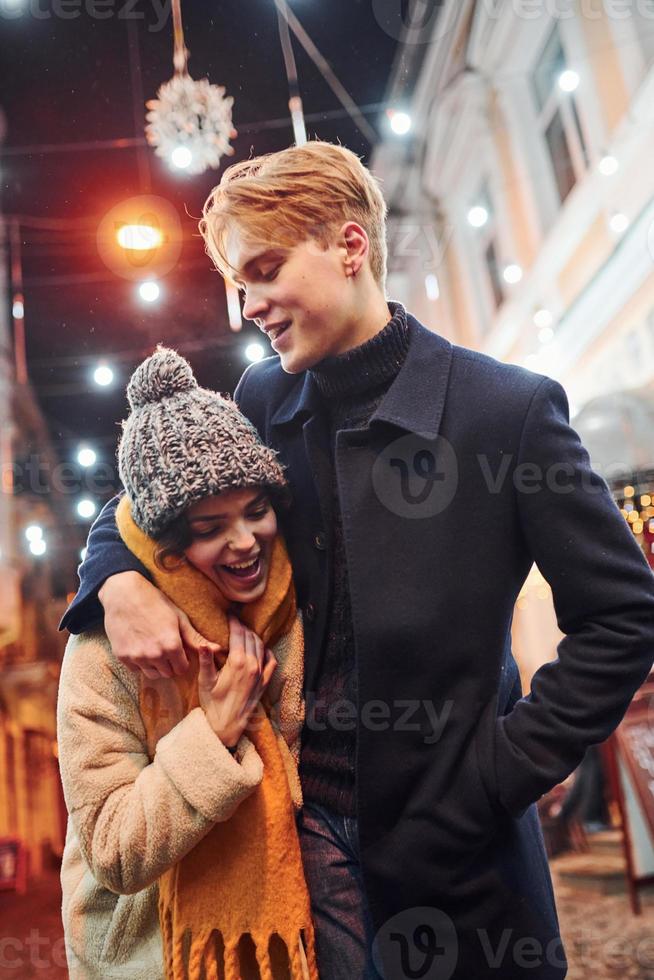 positief jong paar in warm kleren omarmen elk andere Aan Kerstmis versierd straat foto