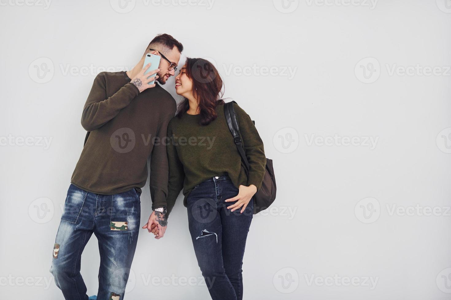vrolijk multi etnisch paar met rugzak en telefoon staand samen binnenshuis in de studio tegen wit achtergrond foto