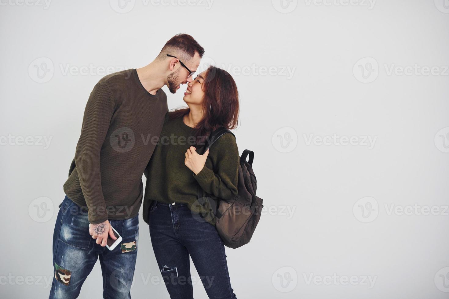 vrolijk multi etnisch paar met rugzak en telefoon staand samen binnenshuis in de studio tegen wit achtergrond foto