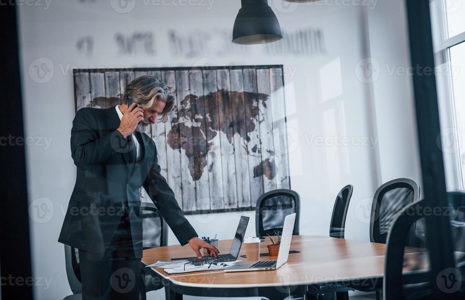 volwassen zakenman met grijs haar- en baard in formeel kleren hebben gesprek door de telefoon in de kantoor foto